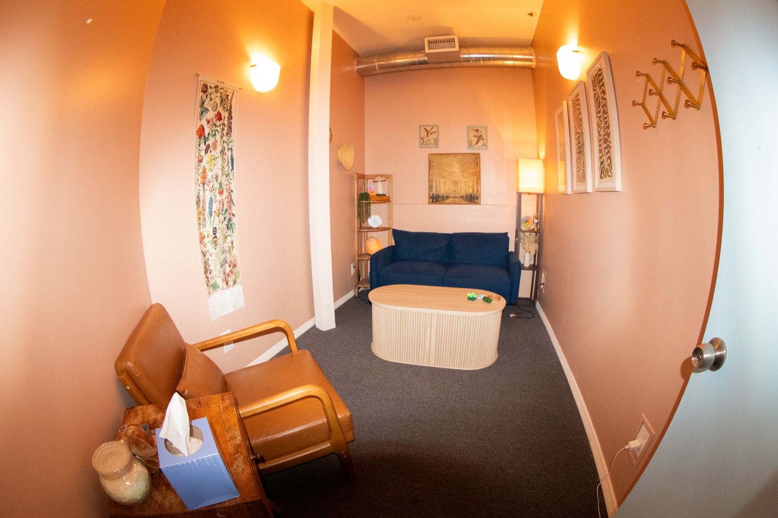 A therapy room with peach walls, a navy blue sofa, a tan leather armchair, a fluted oval coffee table and a botanical wall hanging.